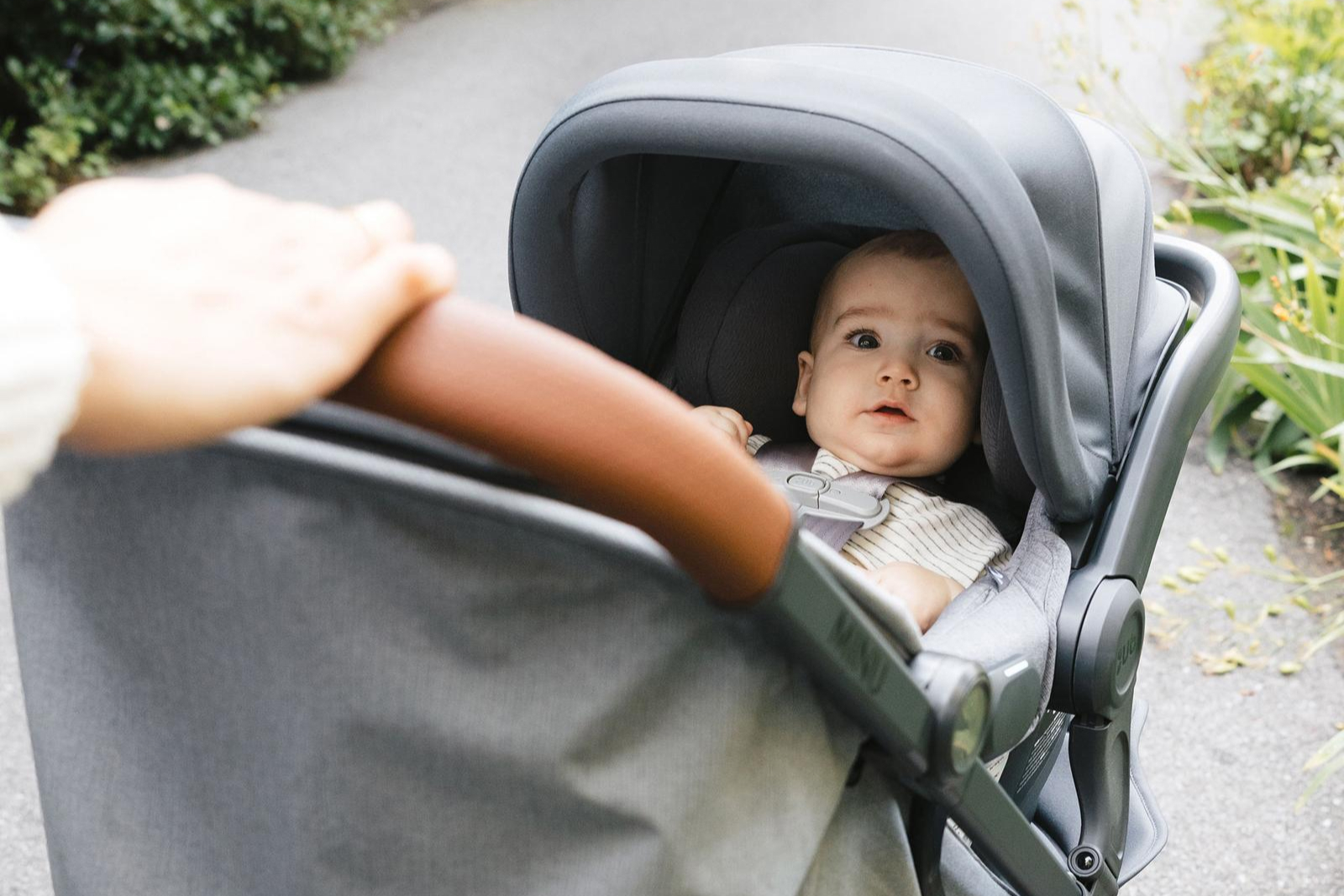 Baby in gray stroller being pushed by adult on outdoor path with greenery in background.