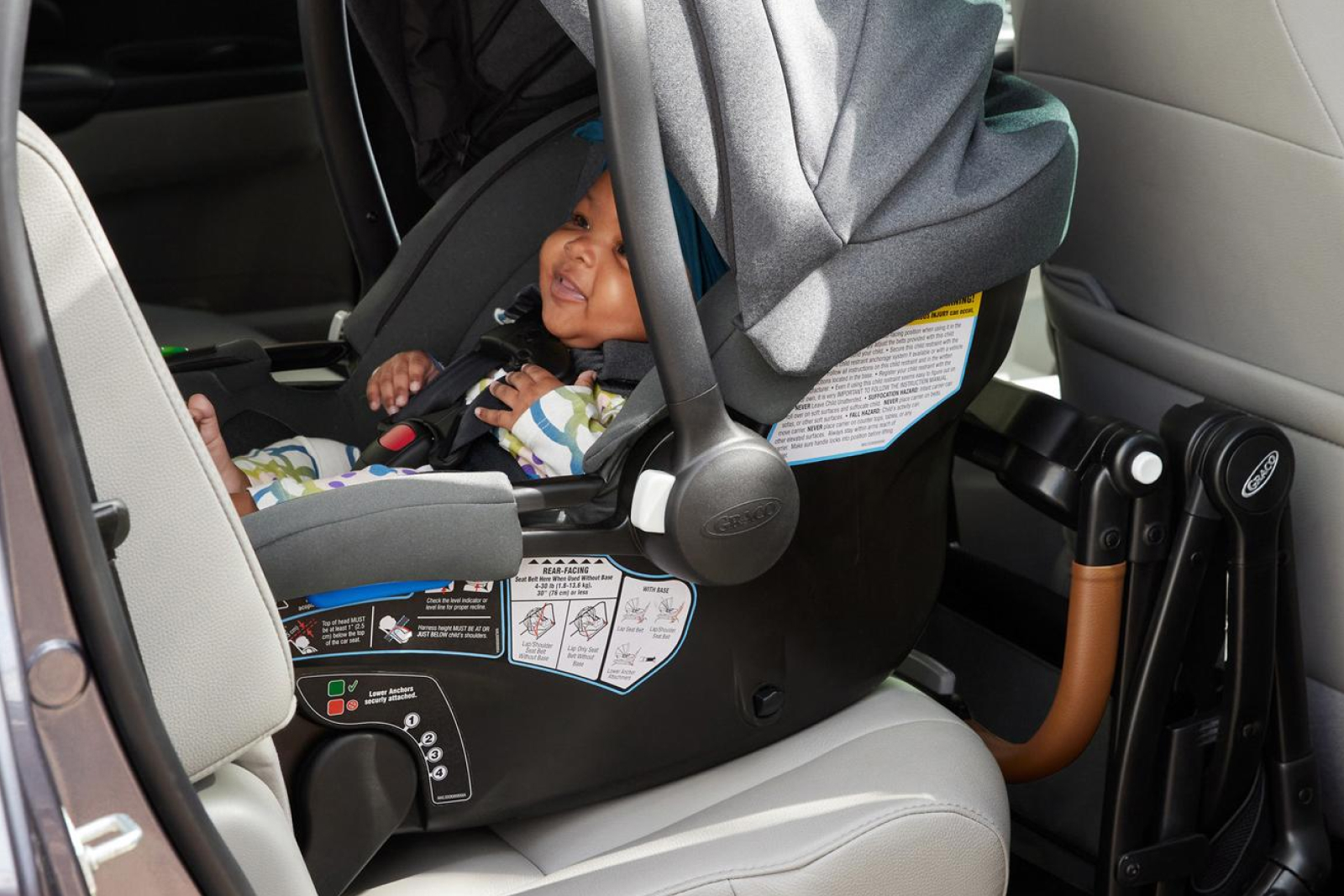 Smiling baby secured in a rear-facing gray car seat with safety harness in vehicle back seat with beige interior.