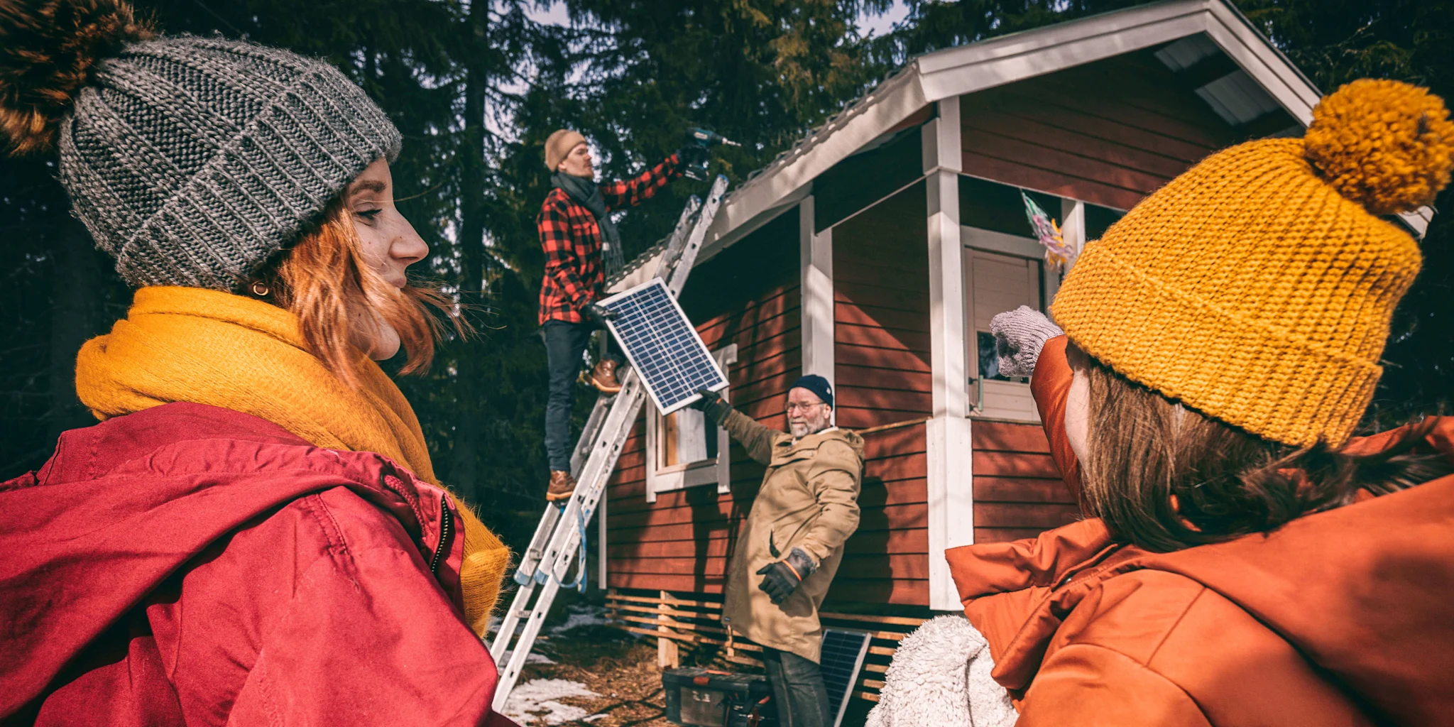 Kaksi naista seisovat etualalla ja tarkkailevat, kun kaksi miestä asentavat aurinkopaneeleja mökin katolle.