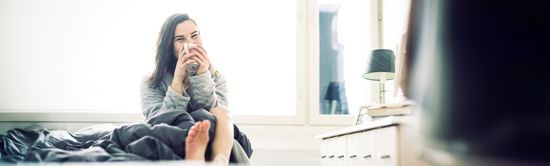 Iloinen nainen istuu sängyn reunalla, hymyilee kameralle ja juo kahvikupista.