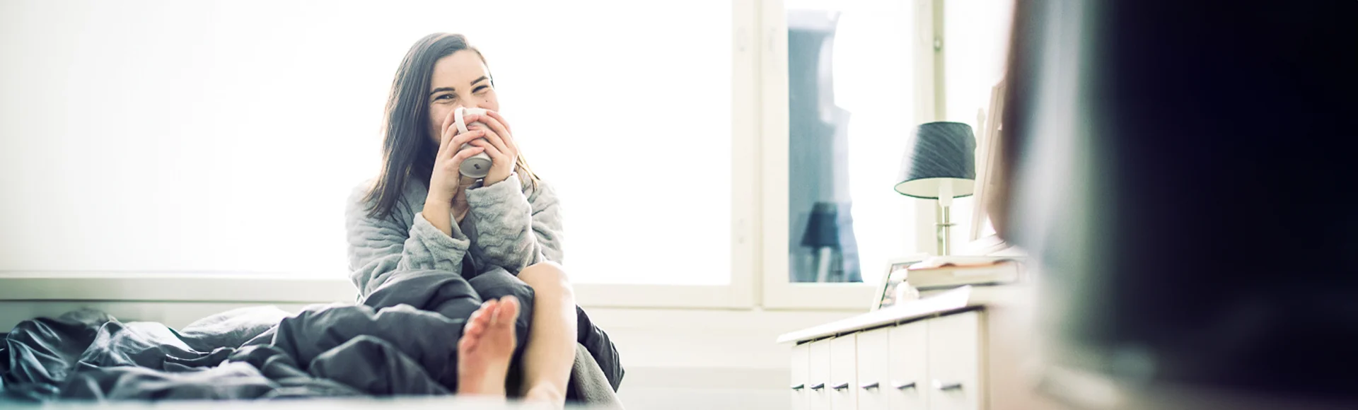 Iloinen nainen istuu sängyn reunalla, hymyilee kameralle ja juo kahvikupista.