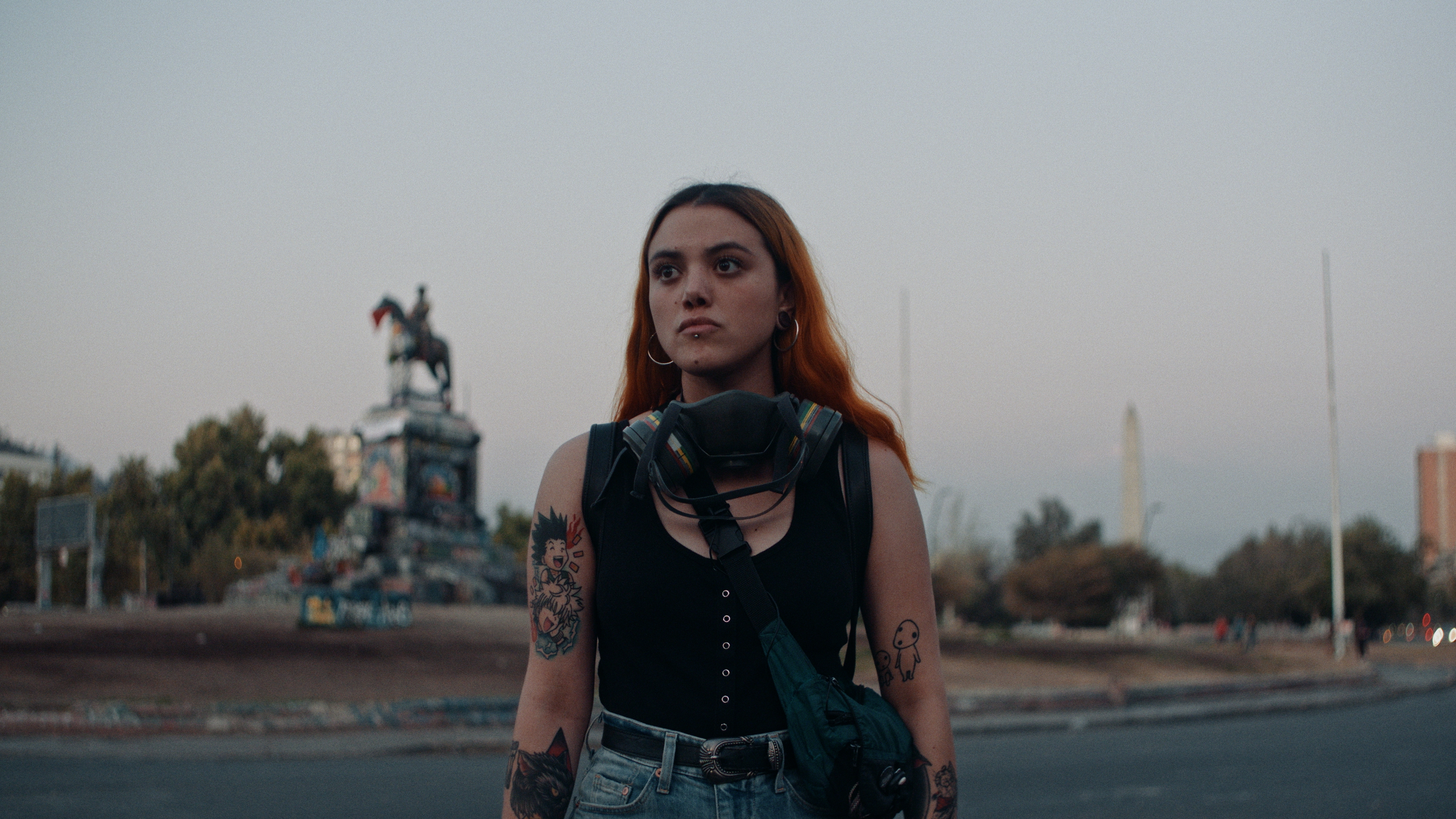 Offizielles Bildmaterial des Films Dear Future Children. Auf dem Bild ist eine junge Frau zu sehen, die auf einer Straße steht. Sie hat Tattoos auf beiden Armen, und eine Gasmaske um ihren Hals. 