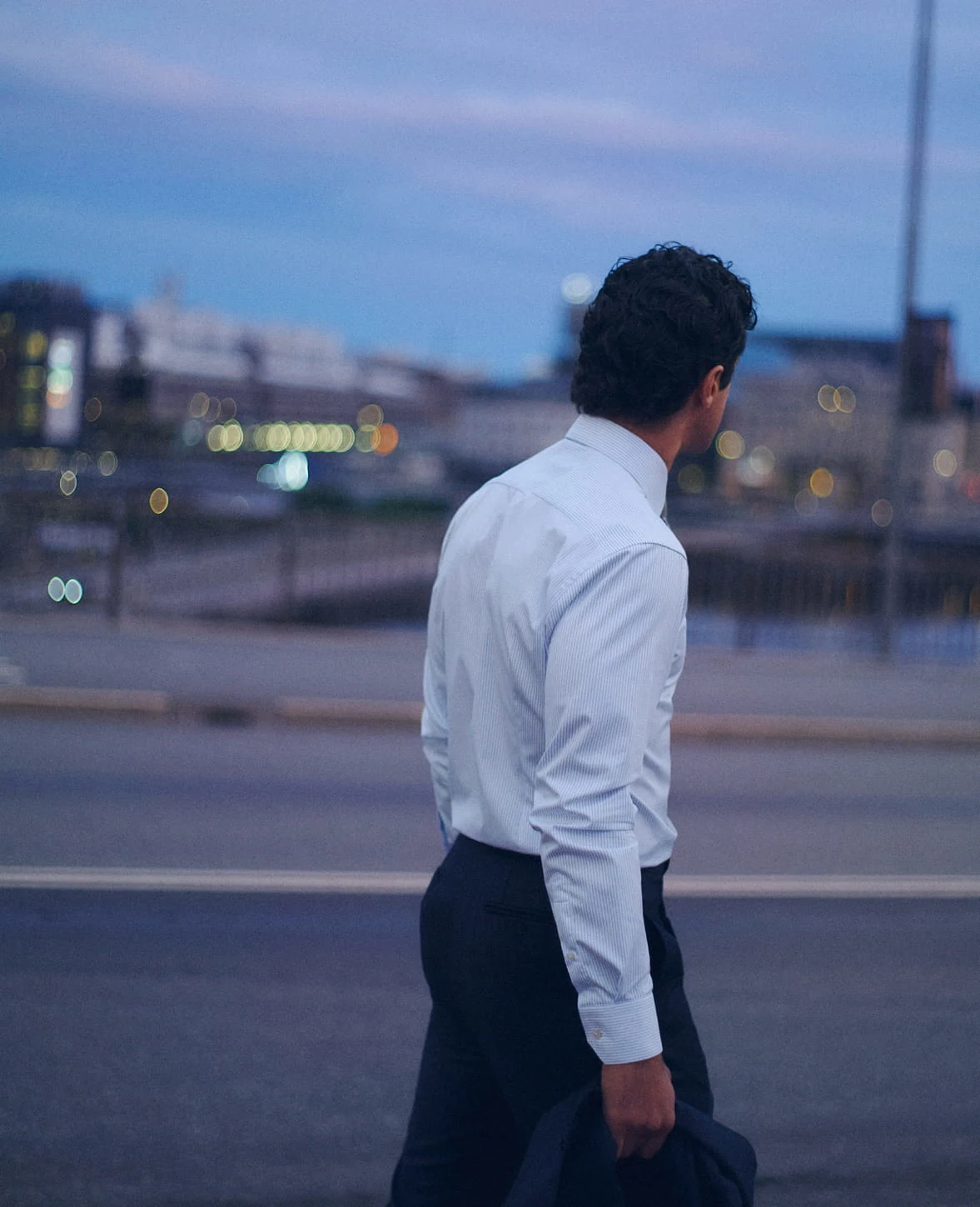 a man walking past an empty street in the evening wearing a tucked in light blue signature twill shirt