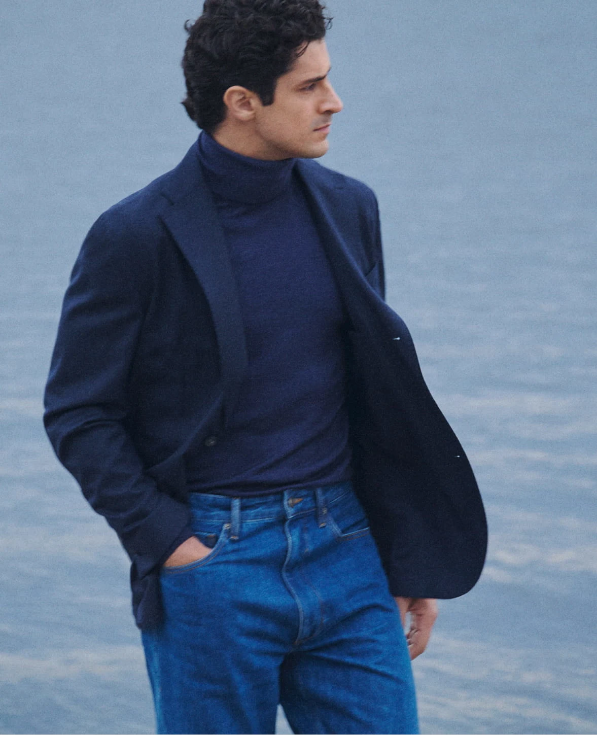 a man posing by the water in a blue turtle neck sweater and a navy soft jacket