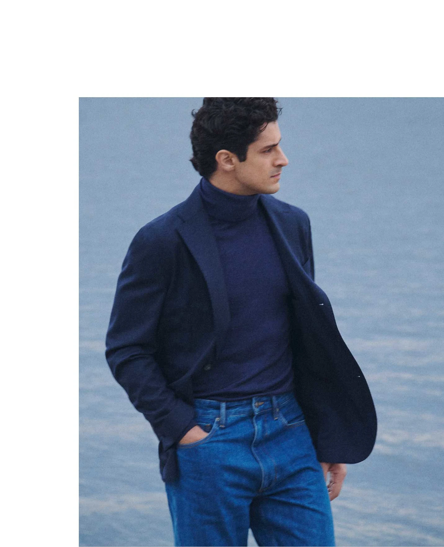 a man posing by the ocean wearing a blue polo sweater and a navy soft jacket