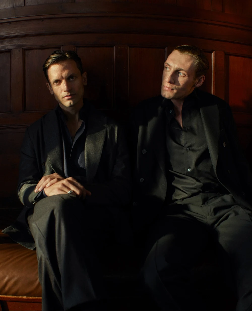 two males sitting by a dark wooden wall with light coming from the side