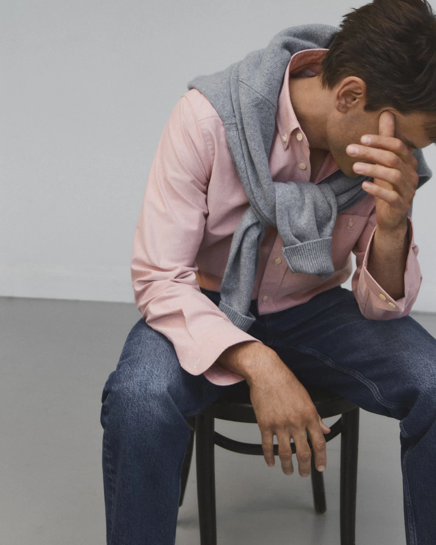 a man posing on a chair in a pink oxford shirt and an knitted crewneck over his shoulders