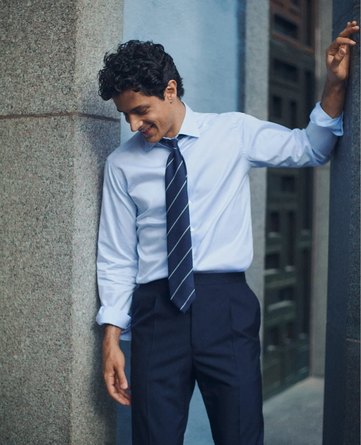 a man leaning against a stone wall in a light blue signature twill shirt and a striped navy grenadine tie