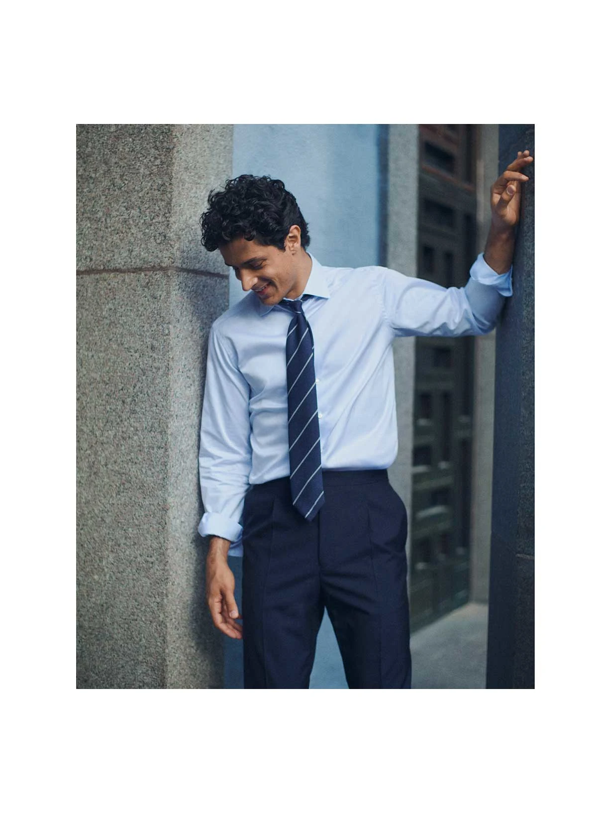 a man leaning against a stone wall in a light blue signature twill shirt and a striped navy grenadine tie