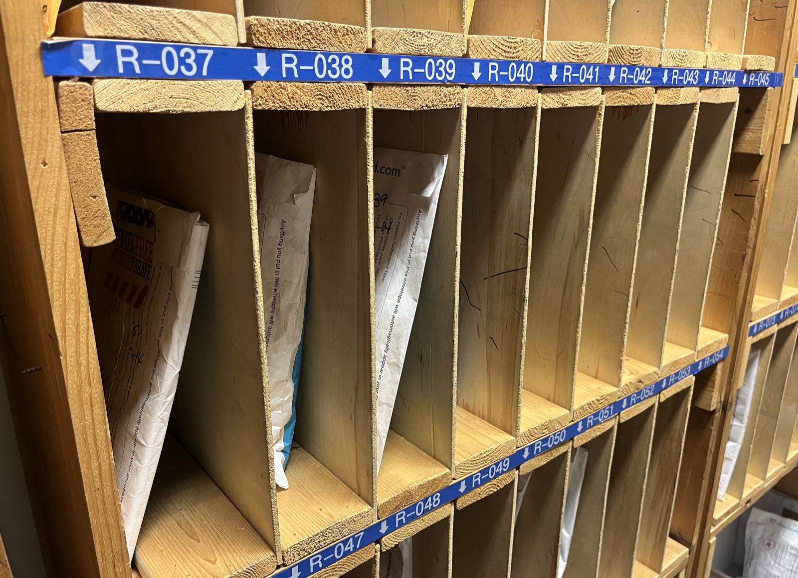 Wooden mailroom-style sorting cubbies labeled R-037 through R-049, holding Magic Envelopes from individual customers awaiting processing