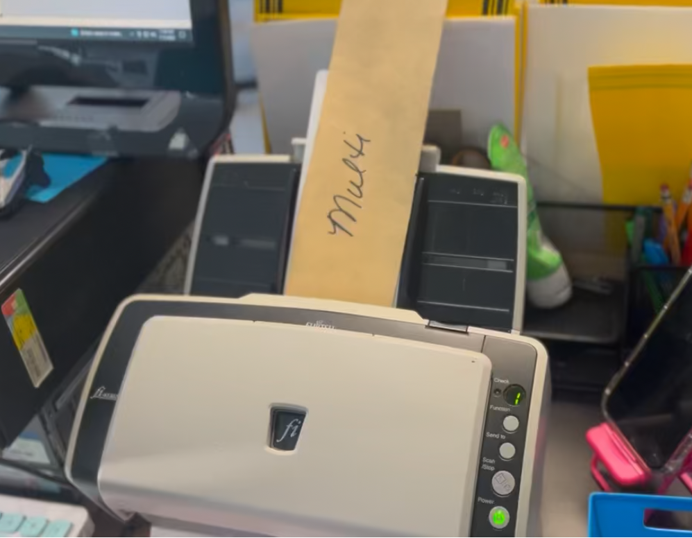 A Shoeboxed Magic Envelope labeled MN H. being fed into a document scanner at the Durham processing station, with a calculator on the desk