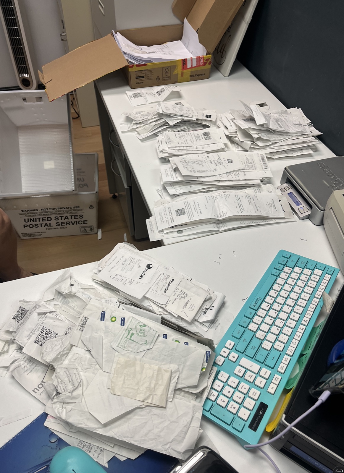 A desk covered with receipts mid-processing, with a USPS mailing box on the floor and a Shoeboxed Magic Envelope being unpacked