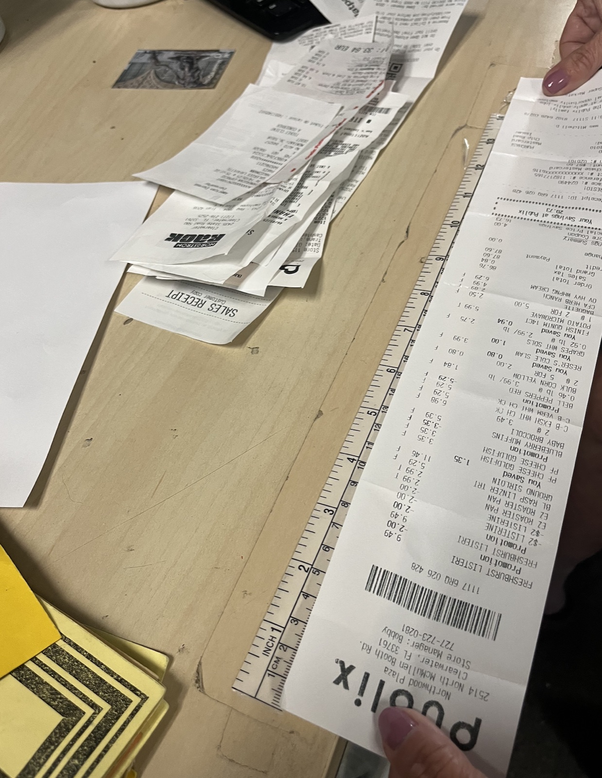 A long Publix grocery receipt held against a 12-inch ruler, showing it stretches well past the ruler's full length, surrounded by other paper receipts on a wooden desk