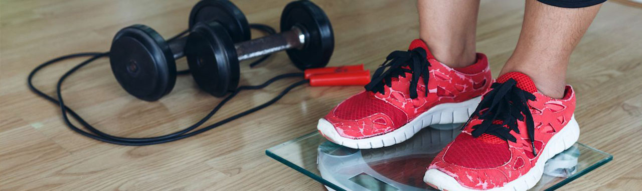 Une femme se pèse avec ses chaussures de sport, à coté d'elle est posé du matériel de fitness