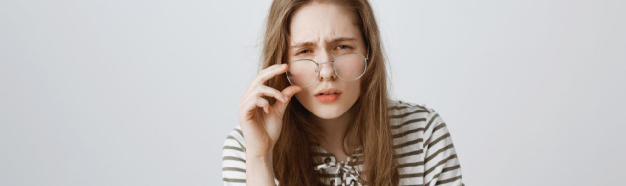 Une femme dos à un mur porte ses lunettes sur le bout de son nez d'un air interrogateur