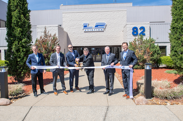 A ribbon cutting outside an office building that says "L2 Aviation" on the front. There are green trees on either side of the building. It is a bright sunny day. Holding the ribbon from left to right is Lee Crume, Be NKY; Kentucky Governor Andy Beshear; Mark Lebovitz, L2 Aviation; Tony Bailey, L2 Aviation; Larry Krauter, CVG; Jeff Rex, L2 Aviation. Tony Bailey is holding a large pair of golden scissors to cut the white ribbon that includes the CVG logo and the L2 Aviation logo and reads April 26, 2025.