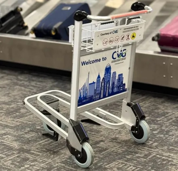 A photo of a luggage cart sitting on a carpeted floor. A baggage carousel with luggage is seen blurred in the background. The cart is facing away from the camera, so you can see the back graphic. The back graphic says "Welcome to CVG" above a graphic of the Cincinnati and Northern Kentucky skyline.