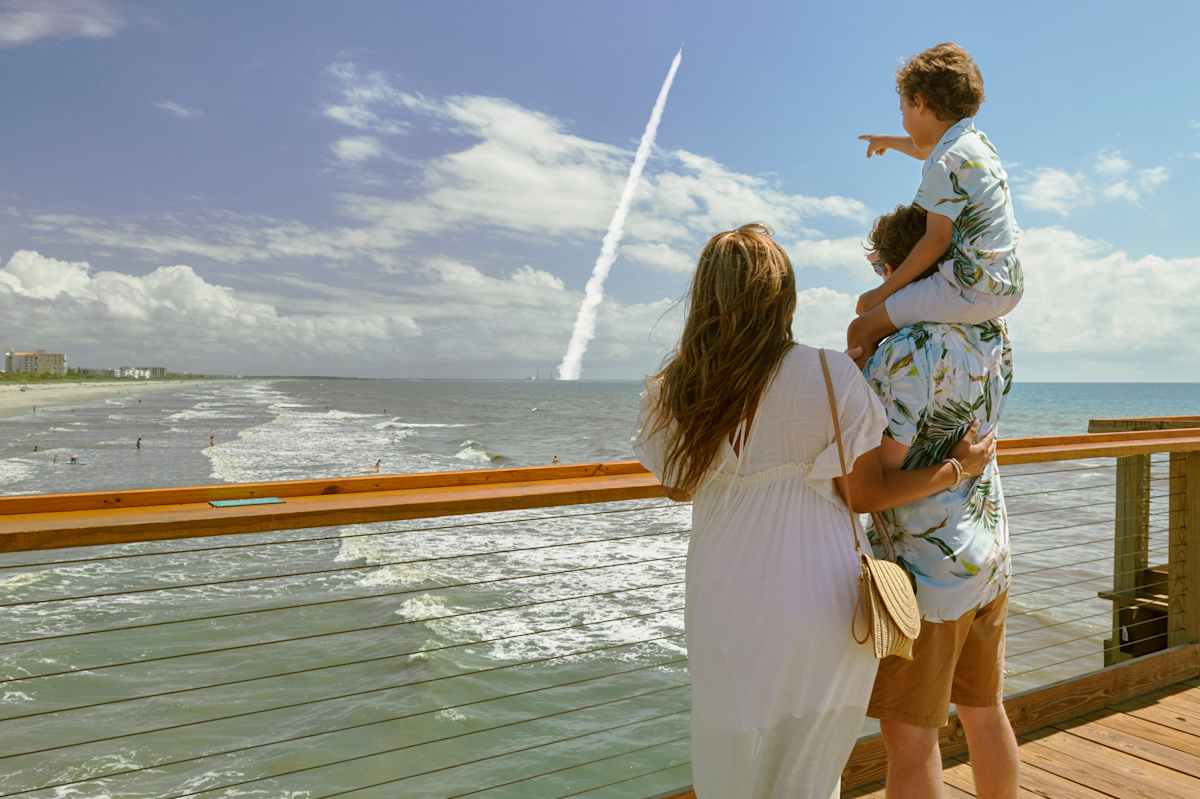 A photo taken from behind of a woman and a man with a child on his shoulders pointing at a rocket taking off in the distance. They are all standing on a pier with the ocean beneath.