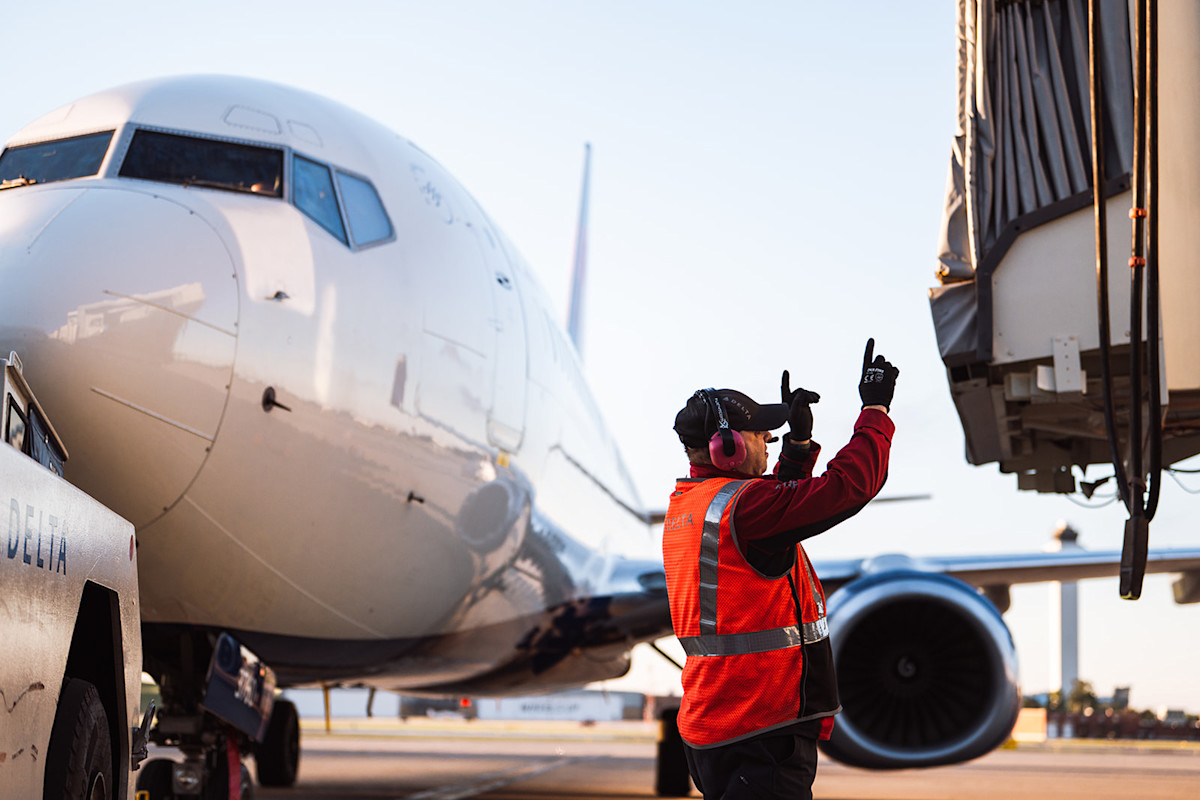A photo of a man dressed in a safety vest with his hands up to direct plane movement. Behind him is a Delta plane attached to a tug with a disconnected jet bridge to the right.