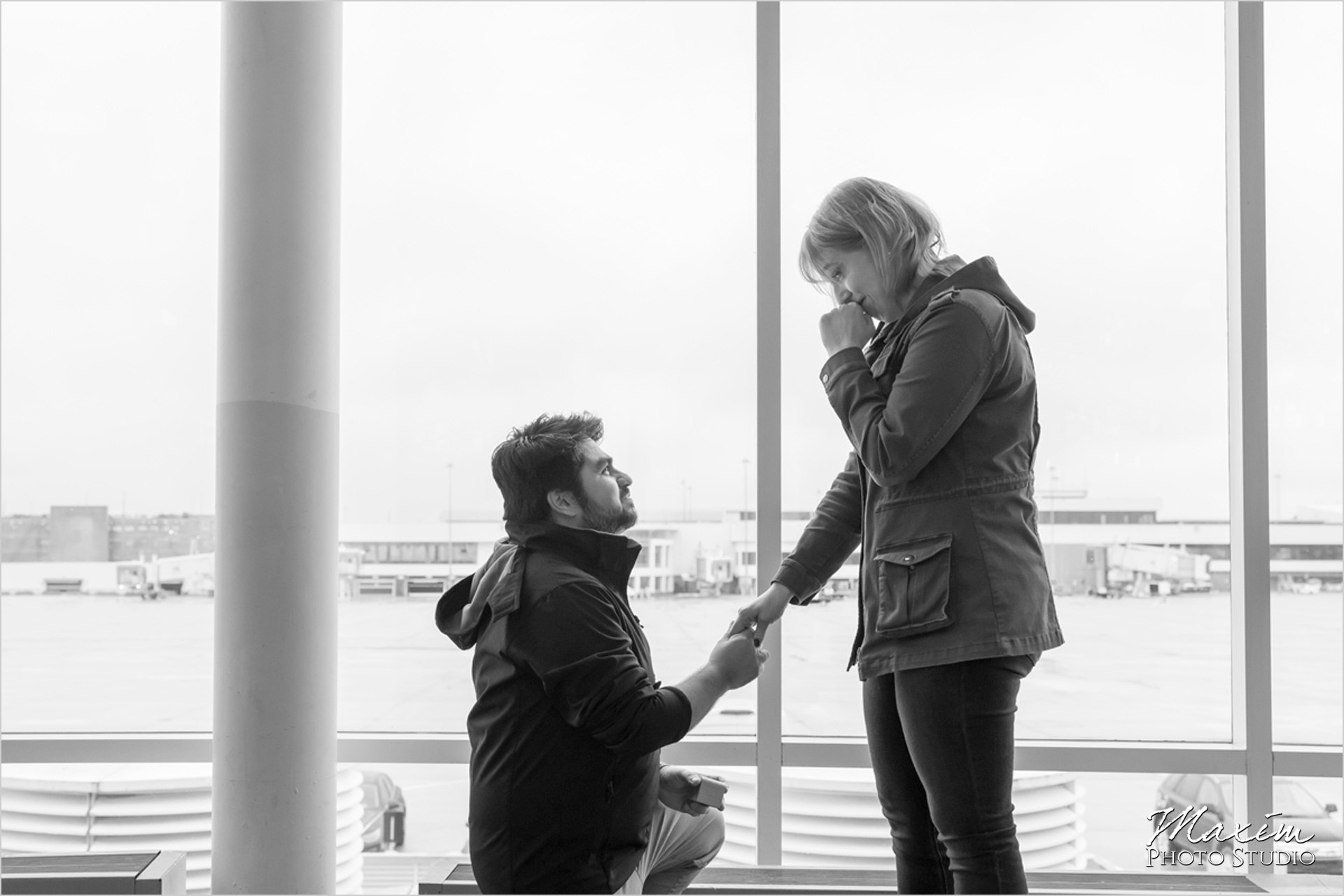 A photo by Maxim Photo Studio. It is a black and white photo of a man on his knee proposing to a woman in front of windows that look out on the airfield.