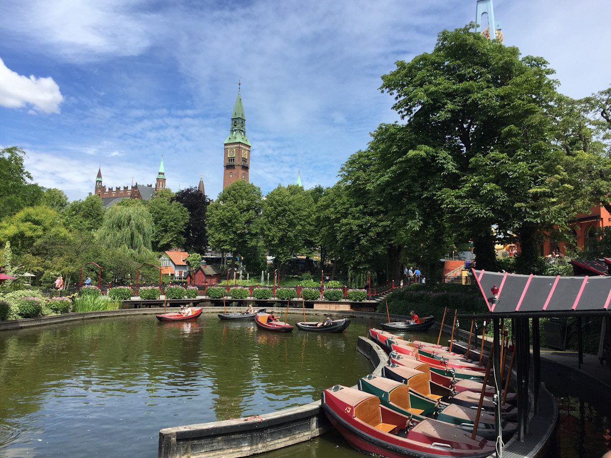 A photo of little row boats on a pond with trees and greenery all around. In the background is a towering building set against a bright blue sky.