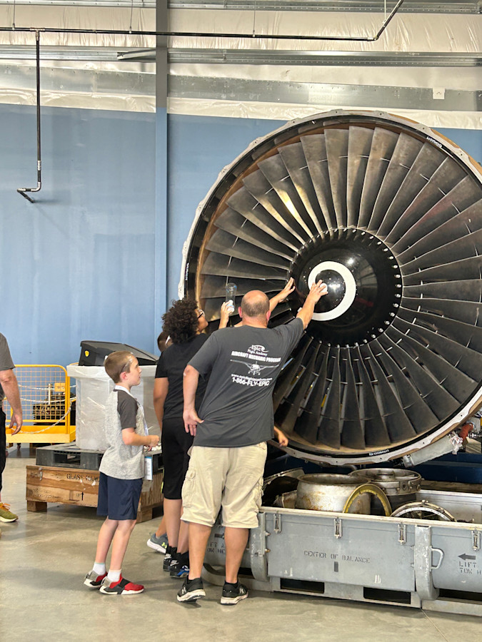 A photo of an adult male and two students touching a stand-alone, stationary plane engine.