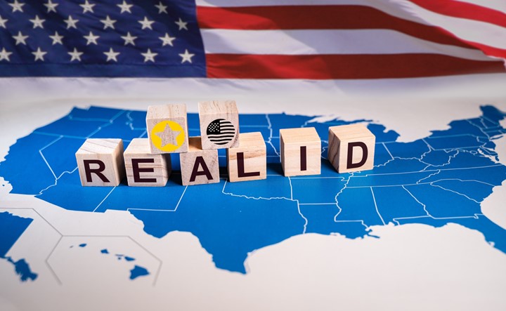 A photo of wooden blocks that spell out Real ID and have a star and an American flag sit on top of a blue map of the United States of America. There is an American flag in the background.