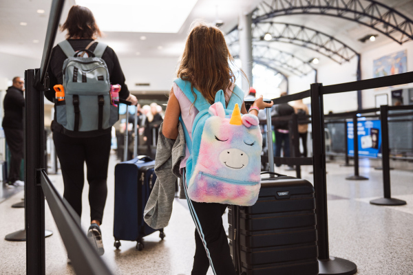 A photo of a little girl in the TSA line with a black suitcase and a unicorn rainbow backpack.