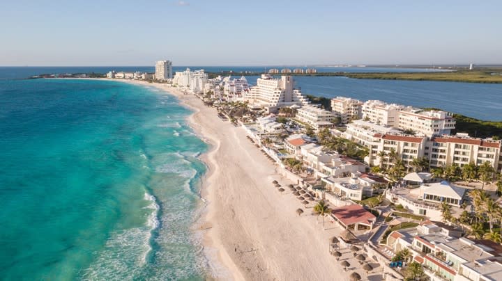 A wide photo of a beach in Cancun with white sand, crystal blue water, and resorts.