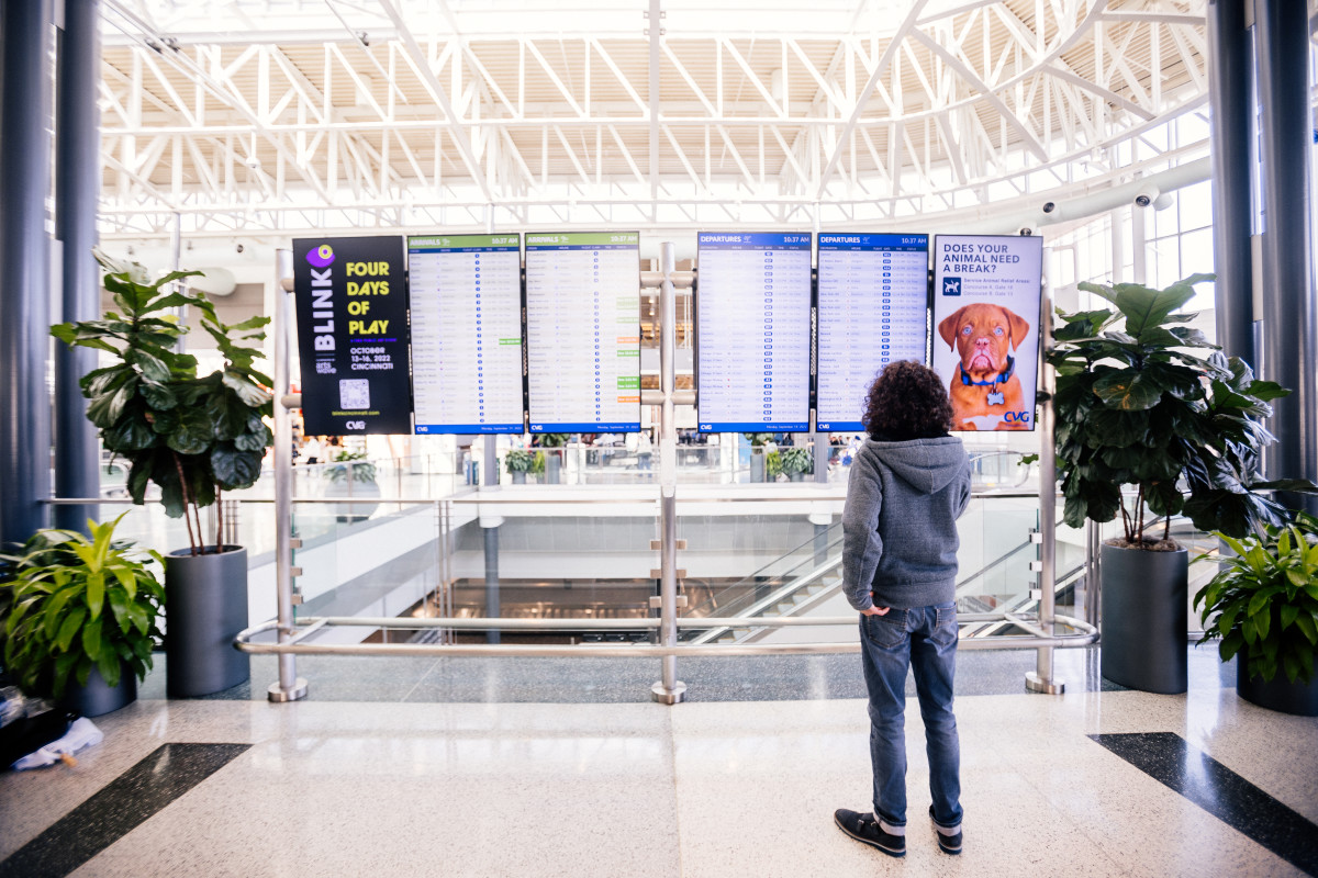 A photo of Jay Kruz standing in front of the Flight Information Display Systems (FIDS) at CVG Airport.