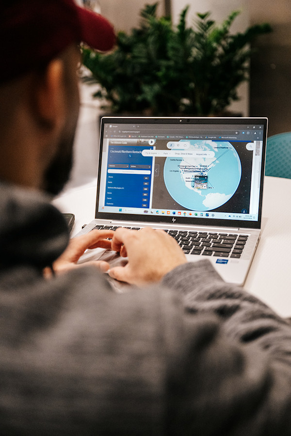 Photo taken from behind a male in a gray sweatshirt with a red baseball cap looking at a laptop with a global map on it.
