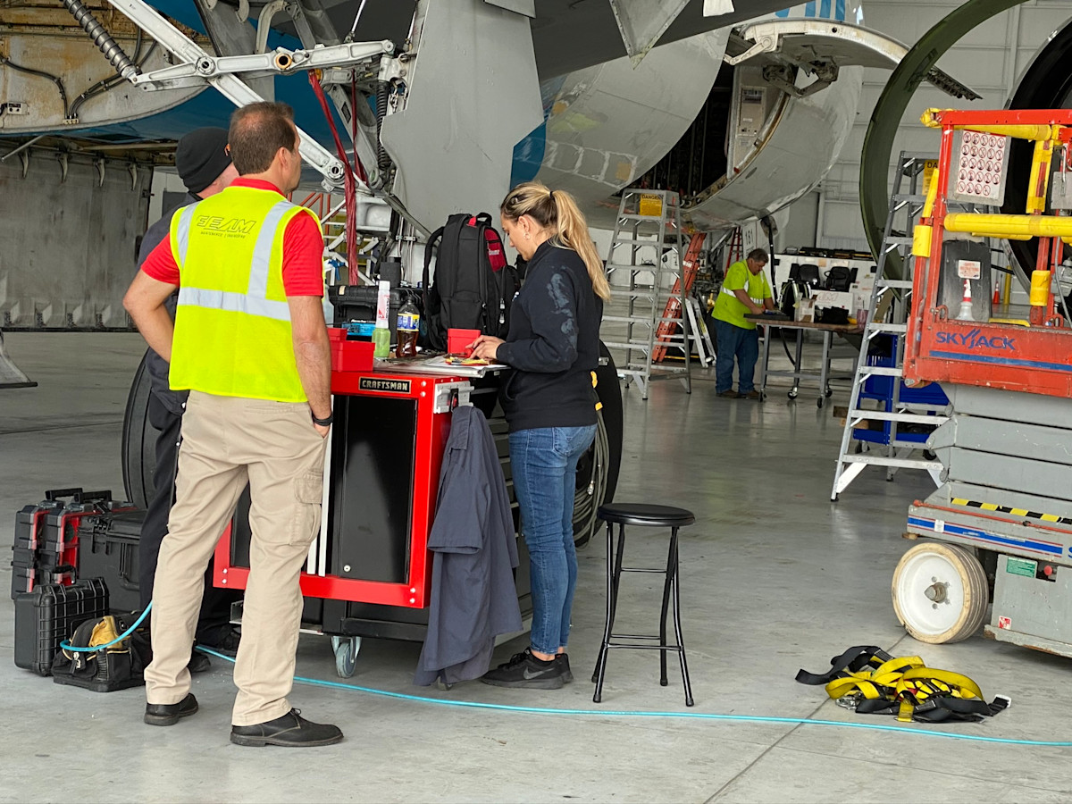 A photo of a male and female aircraft maintenance technician standing at a red cart in an aircraft repair hangar.