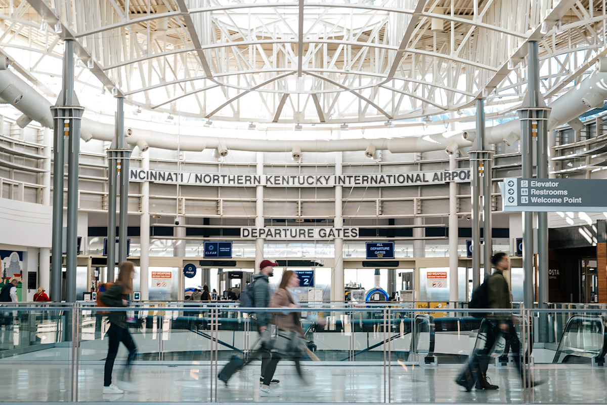 A photo of travelers walking across the Ticketing Bridge at CVG Airport with the TSA checkpoint in the background. The words Cincinnati / Northern Kentucky International Airport and Departure Gates can be seen on the wall in the background.