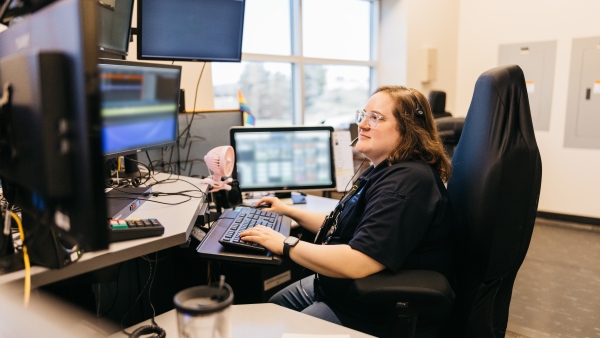 A female telecommunicator sitting in front of her computers and wearing a headset working.