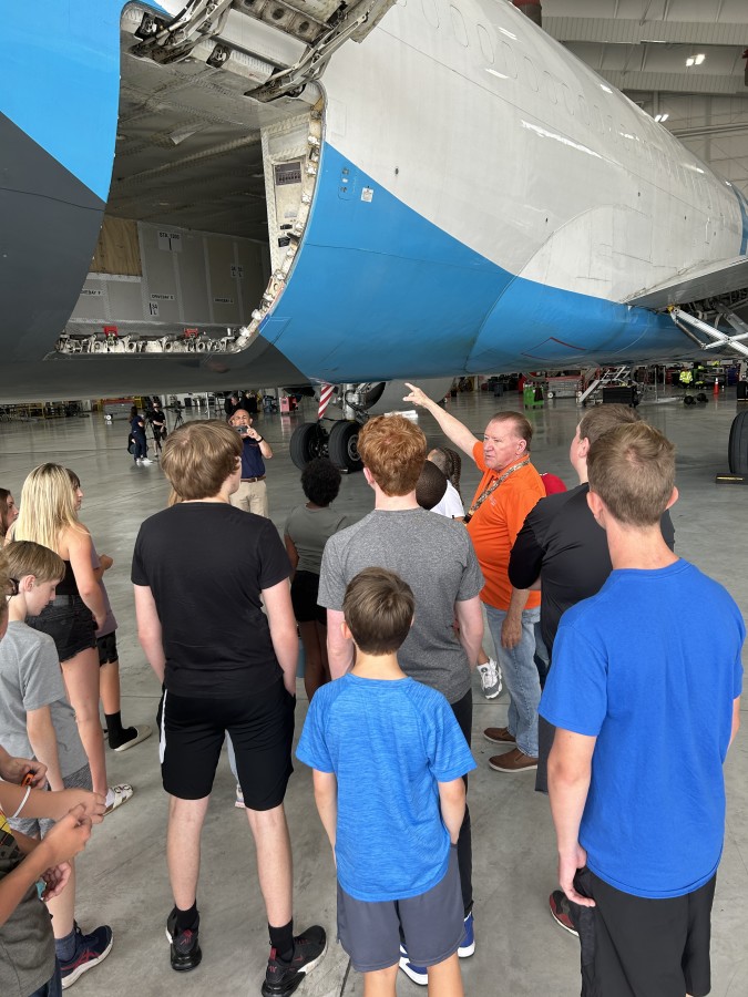 A photo of students on a tour that are standing listening to someone lecture about the plane before them. Their backs are to the camera.