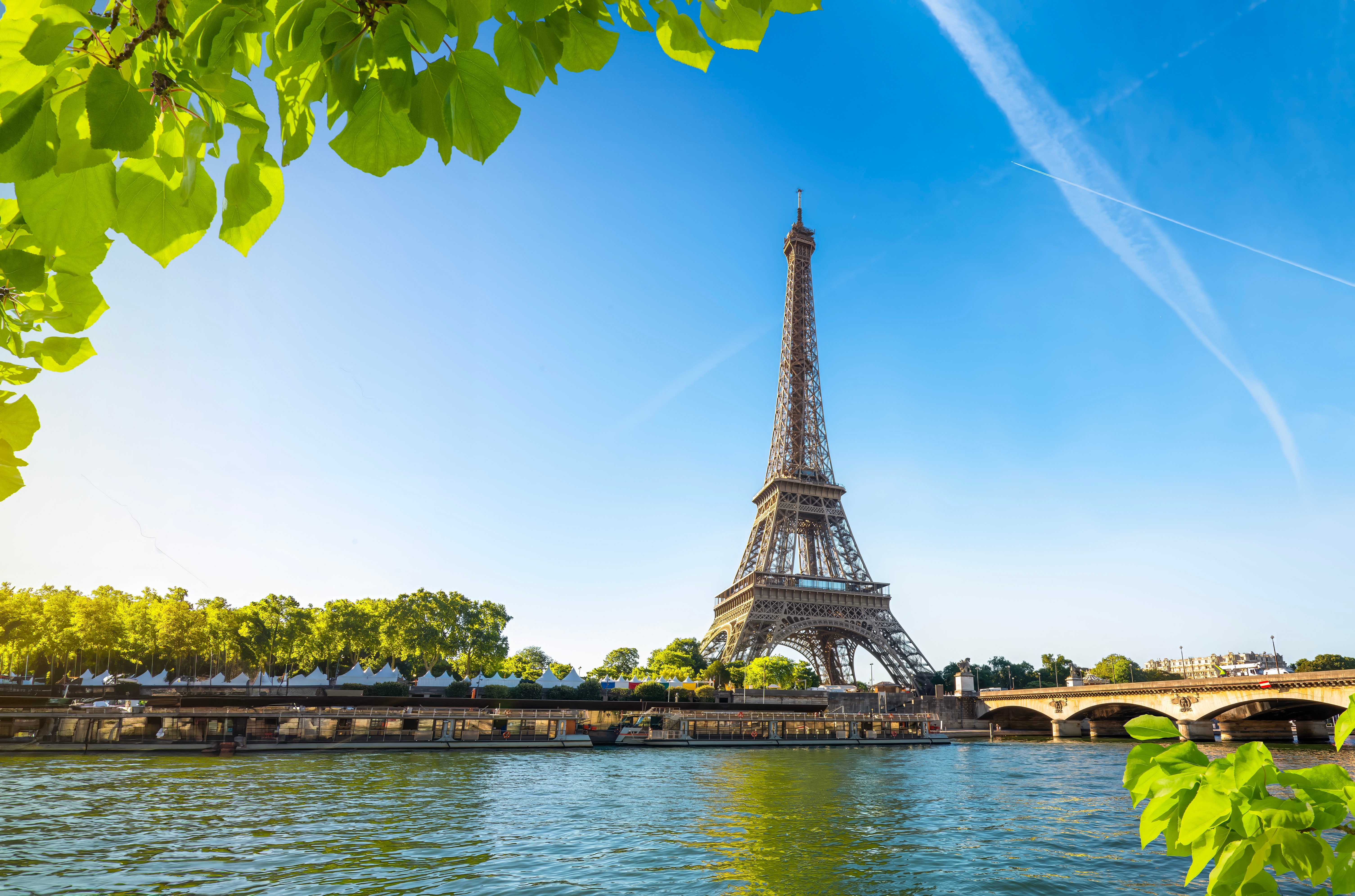 A photo of the Eiffel Tower taken from across the Seine. It is a clear day with a blue sky and greenery around the edges of the photo.