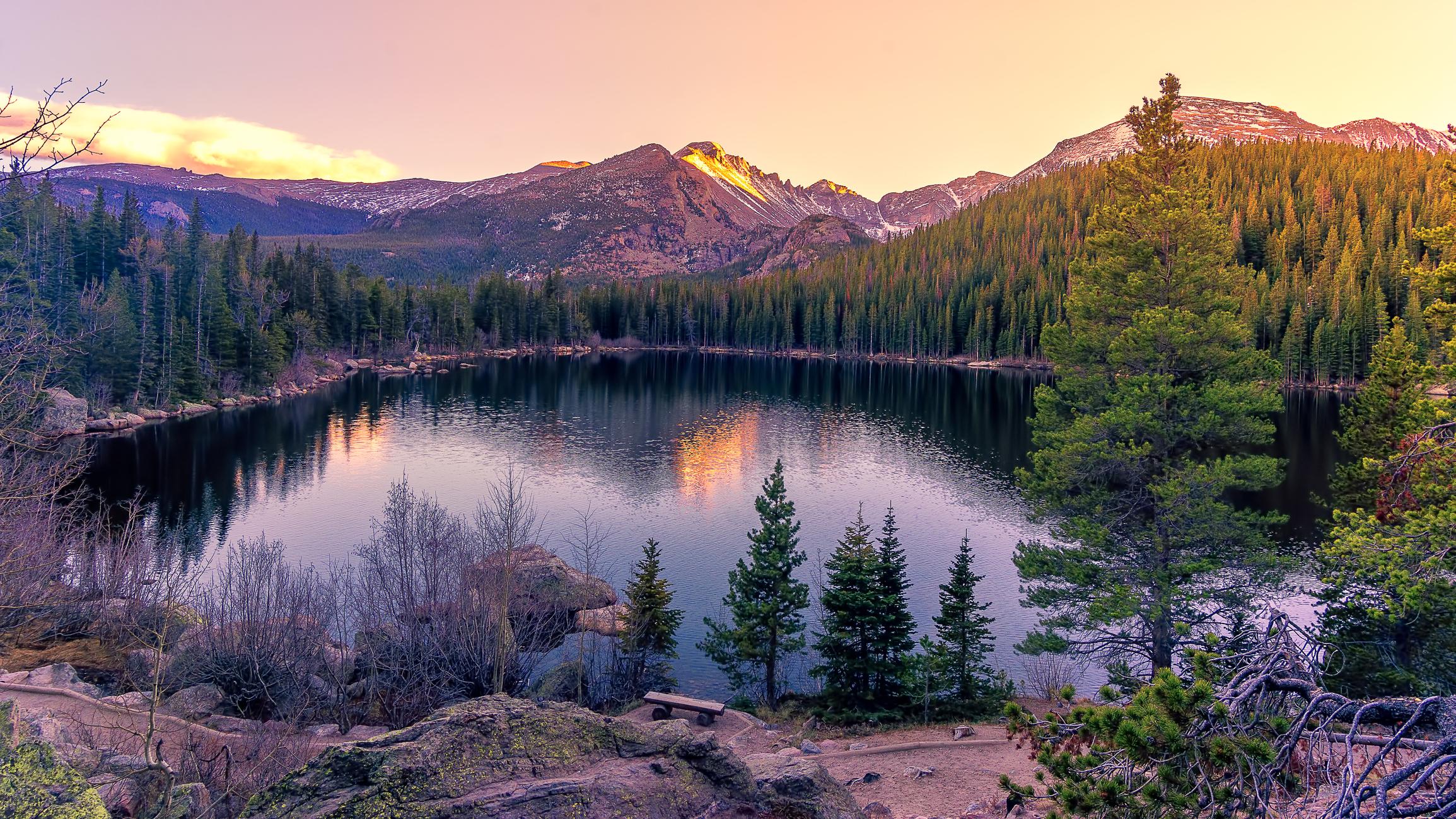 A photo of the Rocky Mountain State Park. There is a lake with mountains and forest in the background with a pink and yellow sky.