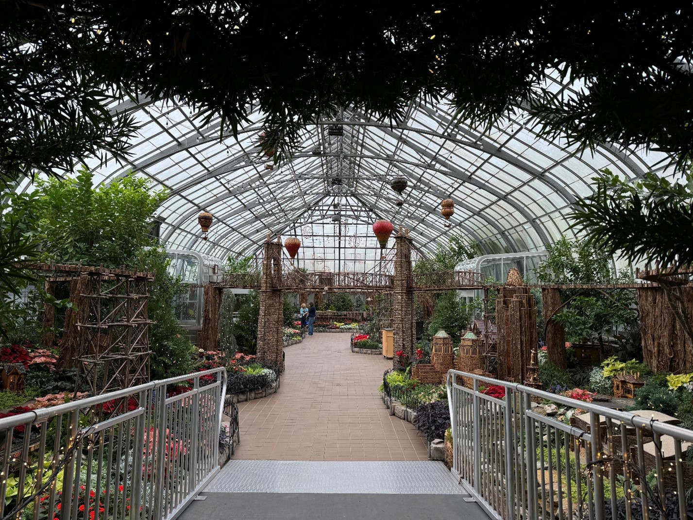 A photo taken from an entrance looking into a room filled with plants and greenery, as well as mini replicas of Cincinnati landmarks made of organic materials. There is a bridge and hot air balloons visible. The environment is bright and inviting.