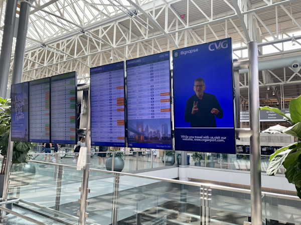 A photo of Flight Information Display Screens in the airport. On one of the screens is a male ASL interpreter.