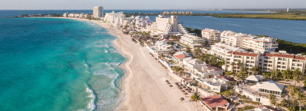 A wide shot photo of a beach in Cancun with the blue water on the left and the building resorts on the right.