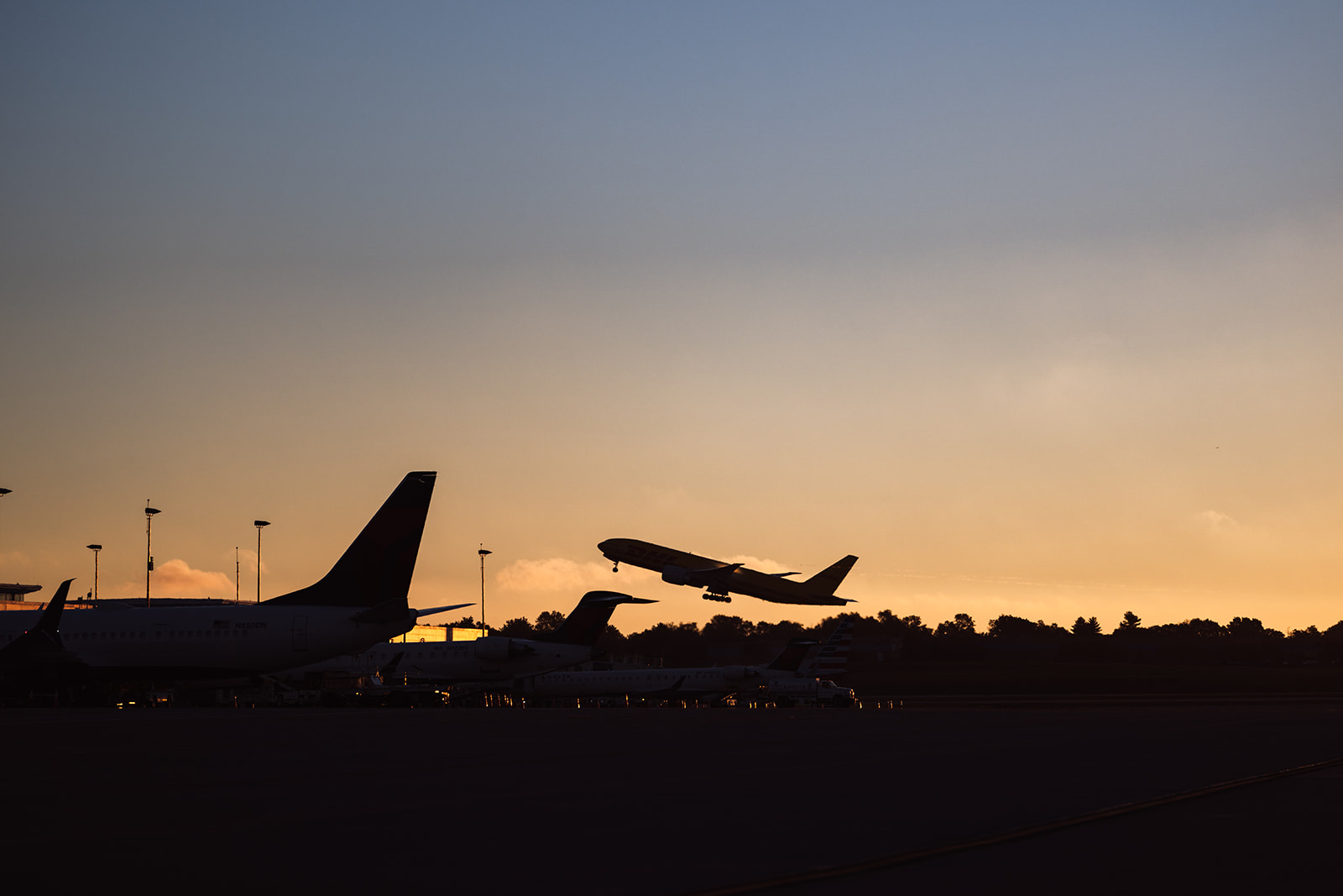 A photo of the silhouette of a plane taking off against a sunset background where the sky is transitioning from blue to orange. Two more planes are visible parked on the ramp.