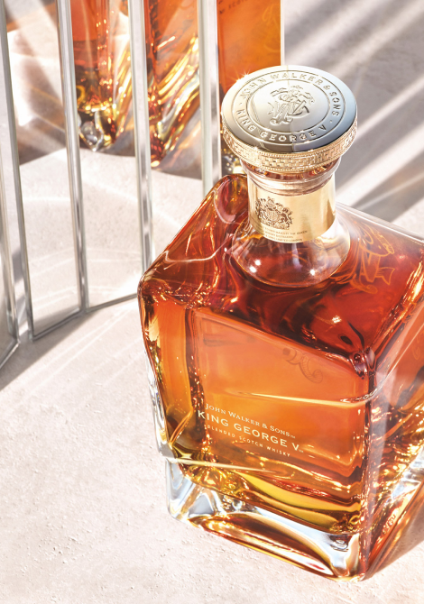 A close-up, top-down shot of a bottle of John Walker & Sons King George V Blended Scotch Whisky, with a soft reflective/ mirror-like background.