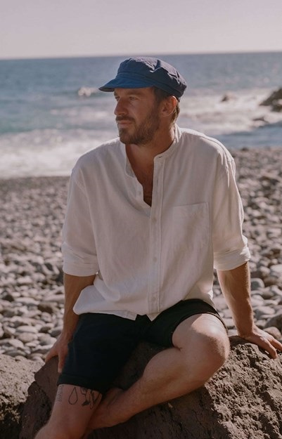 A man wearing a light, long-sleeved shirt and a blue cap is sitting on a rocky beach