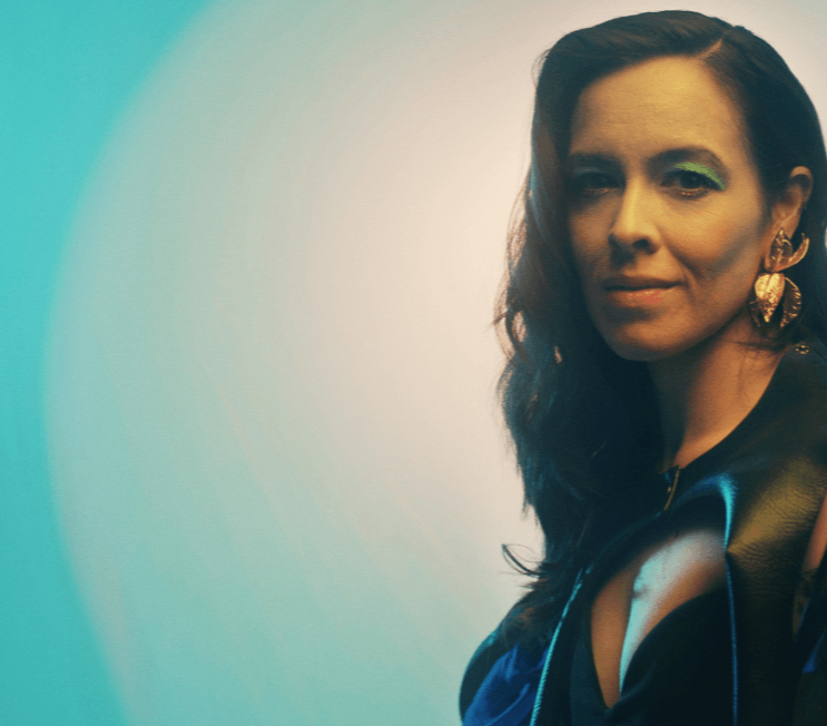 A portrait of a woman with long, dark hair styled in loose waves, wearing bold green eyeshadow and statement gold earrings shaped like leaves.