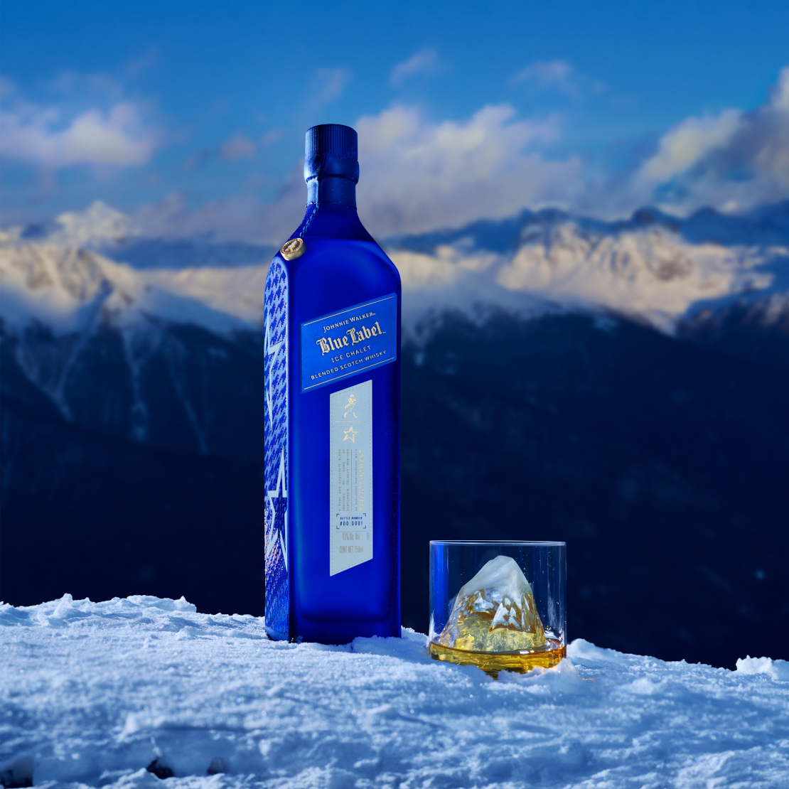 A striking blue bottle of Johnnie Walker Blue Label Ice Chalet stands elegantly on a snow-covered surface with a mountainous backdrop