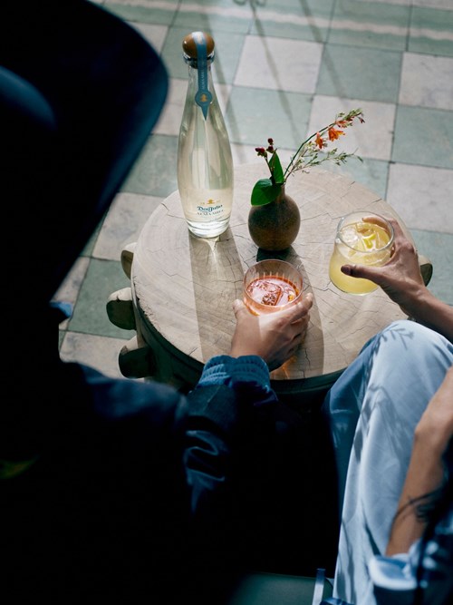 A person’s hand is seen holding a glass with a cocktail on a small, rustic wooden table.