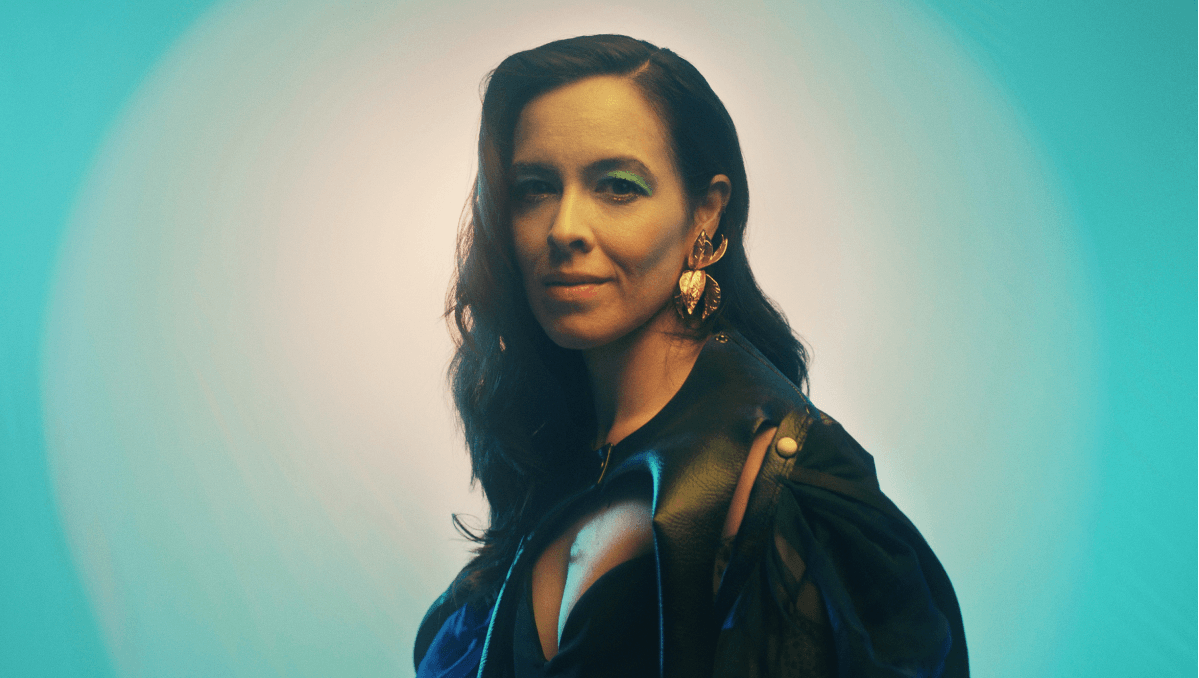 A portrait of a woman with long, dark hair styled in loose waves, wearing bold green eyeshadow and statement gold earrings shaped like leaves.