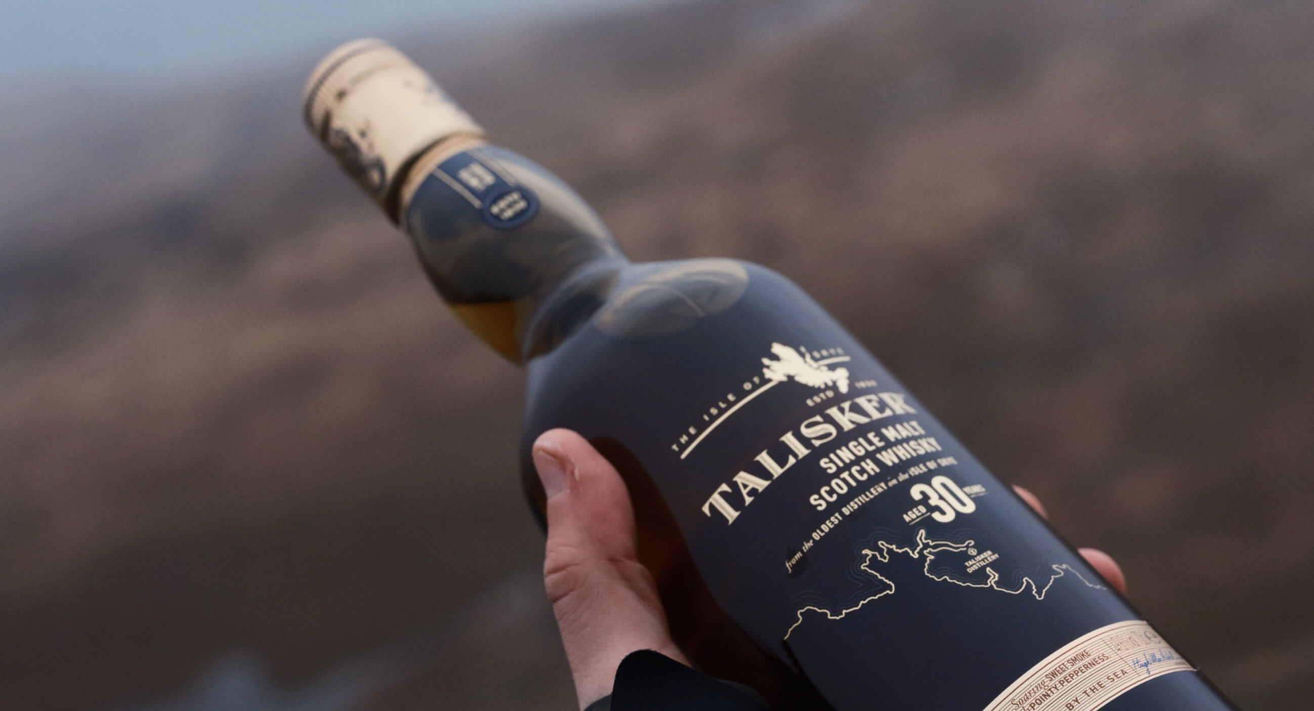 The image shows a close-up of a hand holding a bottle of Talisker 30-Year-Old Single Malt Scotch Whisky against a blurred, earthy background.