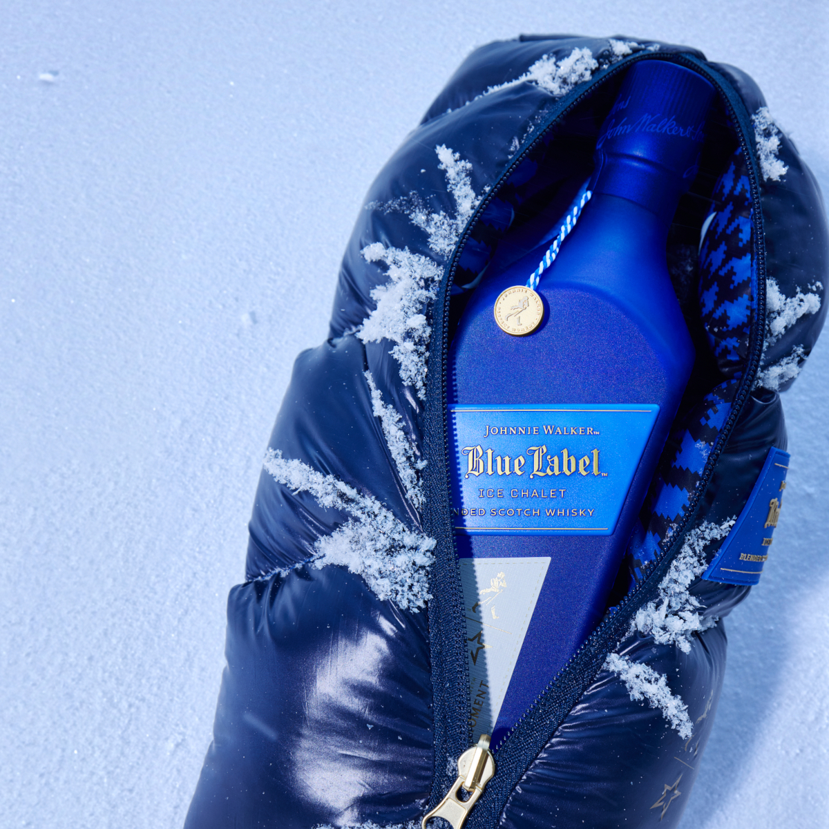 A bottle of Johnnie Walker Blue Label Ice Chalet nestled inside a luxurious, navy-blue quilted pouch, lightly dusted with snowflakes. 