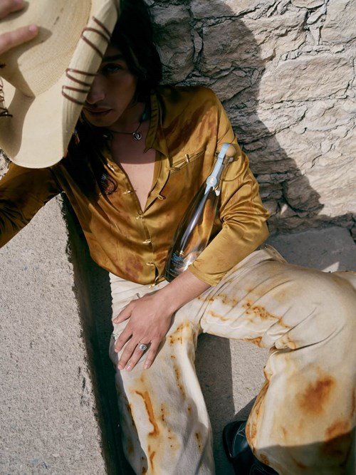 This image displays a woman dressed in a mustard-colored satin shirt and light trousers sits against a stone wall,